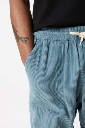 A close-up of a person wearing the light blue Mr Simple Burbank Cord Shorts, featuring an elastic waistband and a white drawstring. The person has a tattoo of a branch on their forearm and is wearing a black shirt, with their hand partially in the shorts pocket—a perfect look for a casual outing.