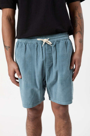 A person wearing light blue Mr Simple Burbank Cord Shorts with a white drawstring stands with their arms by their sides. Sporting a black shirt and visible tattoos on their left forearm, they epitomize the look for a casual outing. The background is plain white.