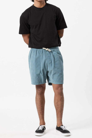 A person is standing against a plain white background. They are wearing a black T-shirt, Mr Simple Burbank Cord Short in blue, and black sneakers with white soles. Their arms are positioned behind their back, perfect for a casual outing.