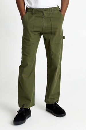 A person stands with hands in pockets, wearing the Mr Simple Carpenter Pant in olive green and black shoes, against a plain white background. Only the lower half of their body is visible.