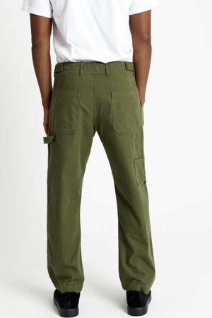 A person faces away from the camera wearing a white T-shirt, black shoes, and the Mr Simple Carpenter Pant in olive green with a workwear-inspired design and large back pockets, standing against a plain white background.