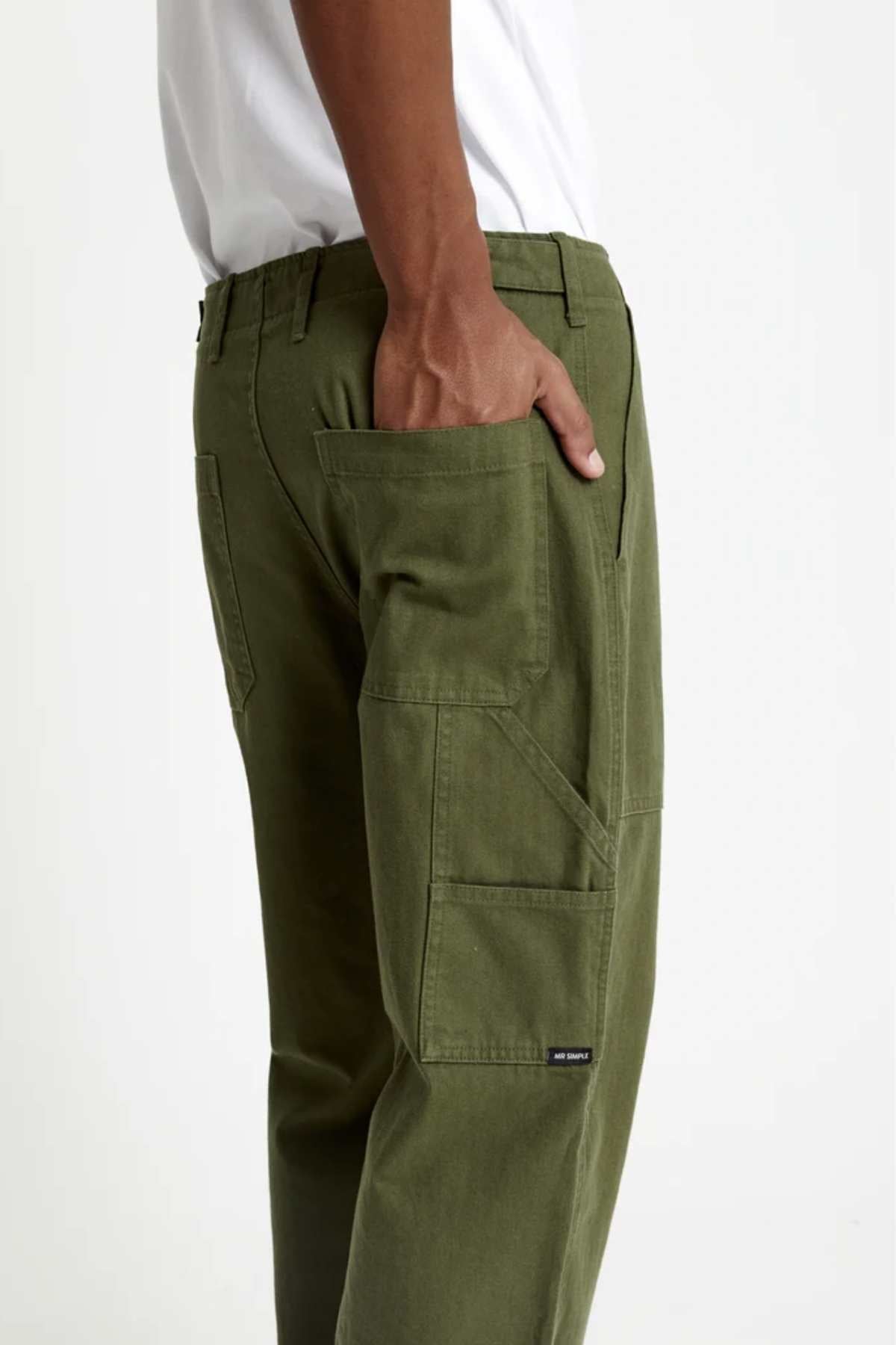 A person wearing the olive green Mr Simple Carpenter Pant, made from 100% cotton with visible stitching and a small black label near the seam, stands with one hand in the side pocket against a plain light background, paired with a white shirt.