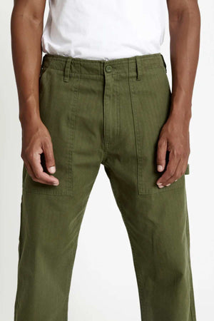 A person wearing the Mr Simple Carpenter Pant in olive green with large front pockets and a white t-shirt, shown from waist to mid-thigh. The workwear-inspired design combines style and functionality.