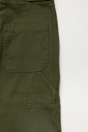 Close-up of the Mr Simple Carpenter Pant in olive green, crafted from 100% cotton with visible stitching, a large rectangular pocket, and a seam detail on the right leg, set against a light background.