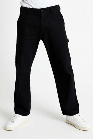 A person wearing relaxed-fit Mr Simple Carpenter Pant and white sneakers stands against a plain white background. The pant features side pockets, while the low-cut sneakers maintain a minimal design, embodying a workwear-inspired aesthetic.
