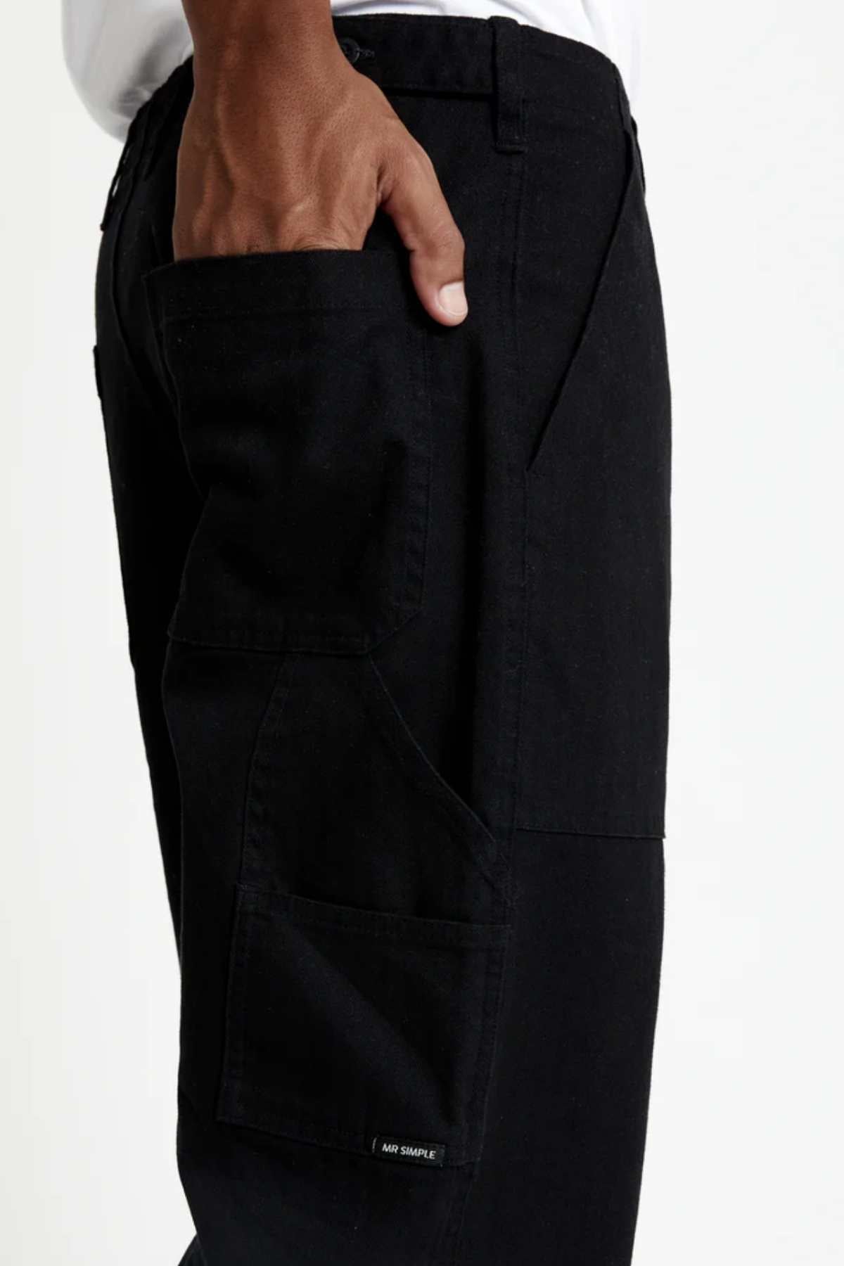 A person wears Mr Simple Carpenter Pants, featuring a relaxed fit and black color. Their hand rests in one of the multiple side pockets, showcasing a workwear-inspired design with a visible logo tag near the seam. They pair it with a white shirt against a plain white background.