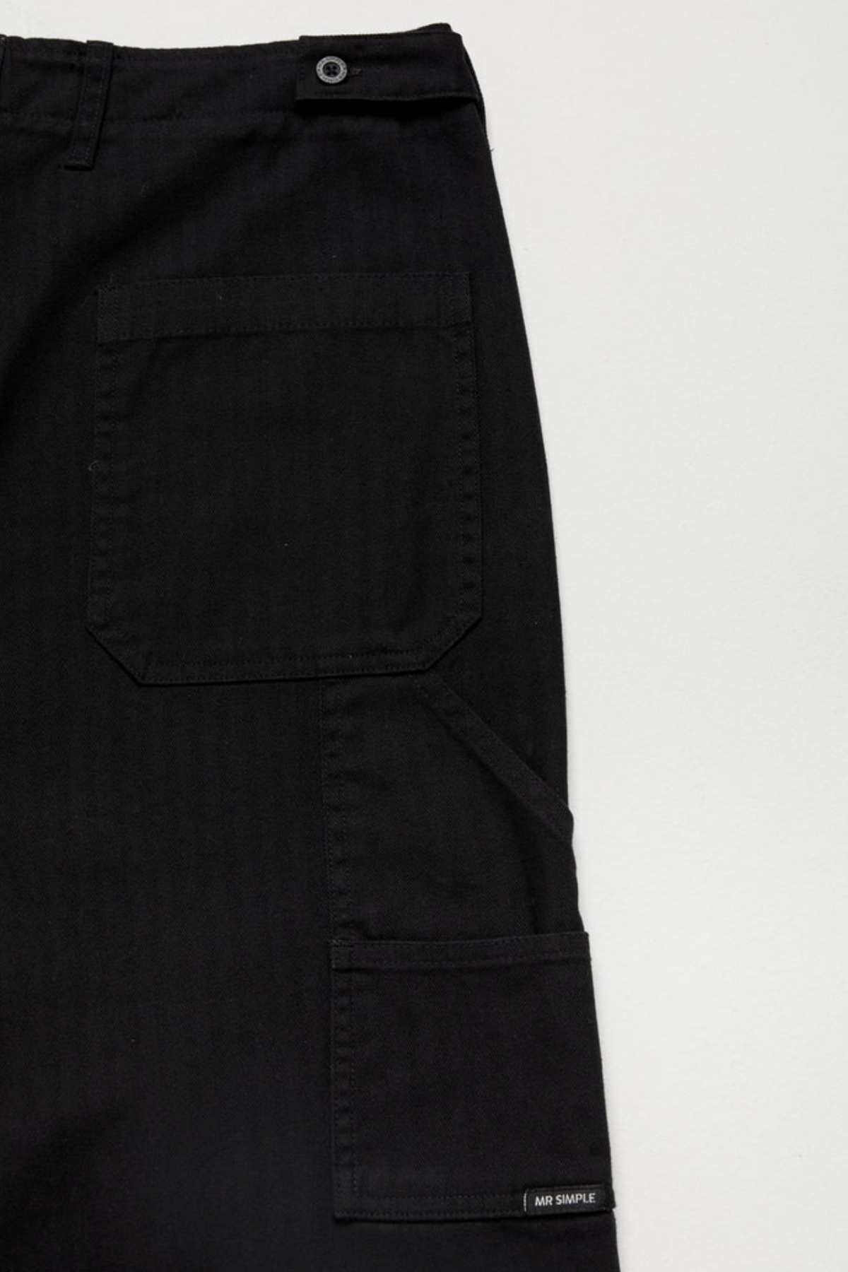 Close-up of the Mr Simple Carpenter Pant in black, showcasing a workwear-inspired design with two pockets and a waist button, set against a plain white background. A small label on the bottom right pocket reads "Aim Simple.