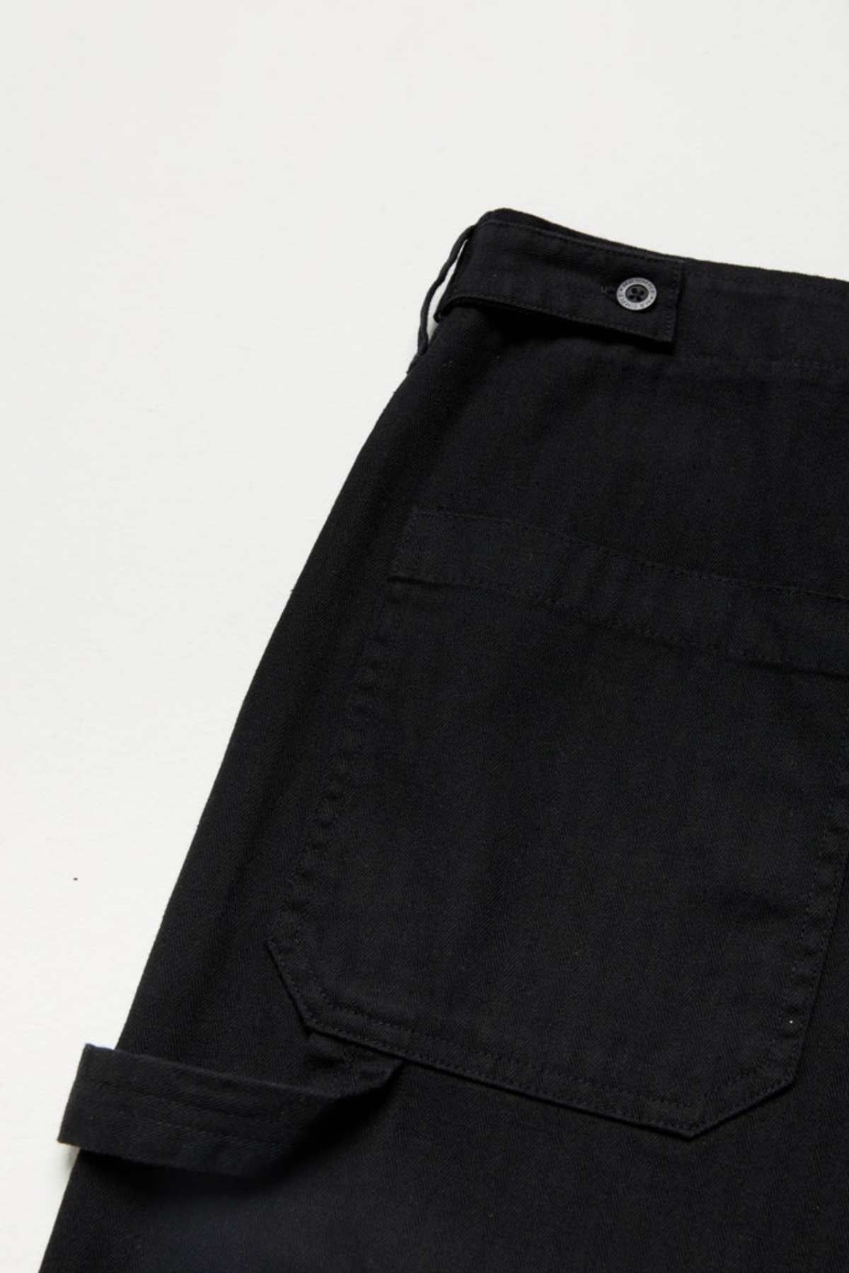 A close-up of the Mr Simple Carpenter Pant shows a pair of black cargo pants with a side pocket, flap, and button details, boasting a relaxed fit and workwear-inspired design. They're displayed on a light-colored surface.