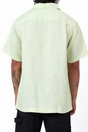A person with tattooed arms is shown from the back, wearing the Mr Simple Cuban Linen Short Sleeve Shirt in light green paired with black pants, standing against a plain white background.