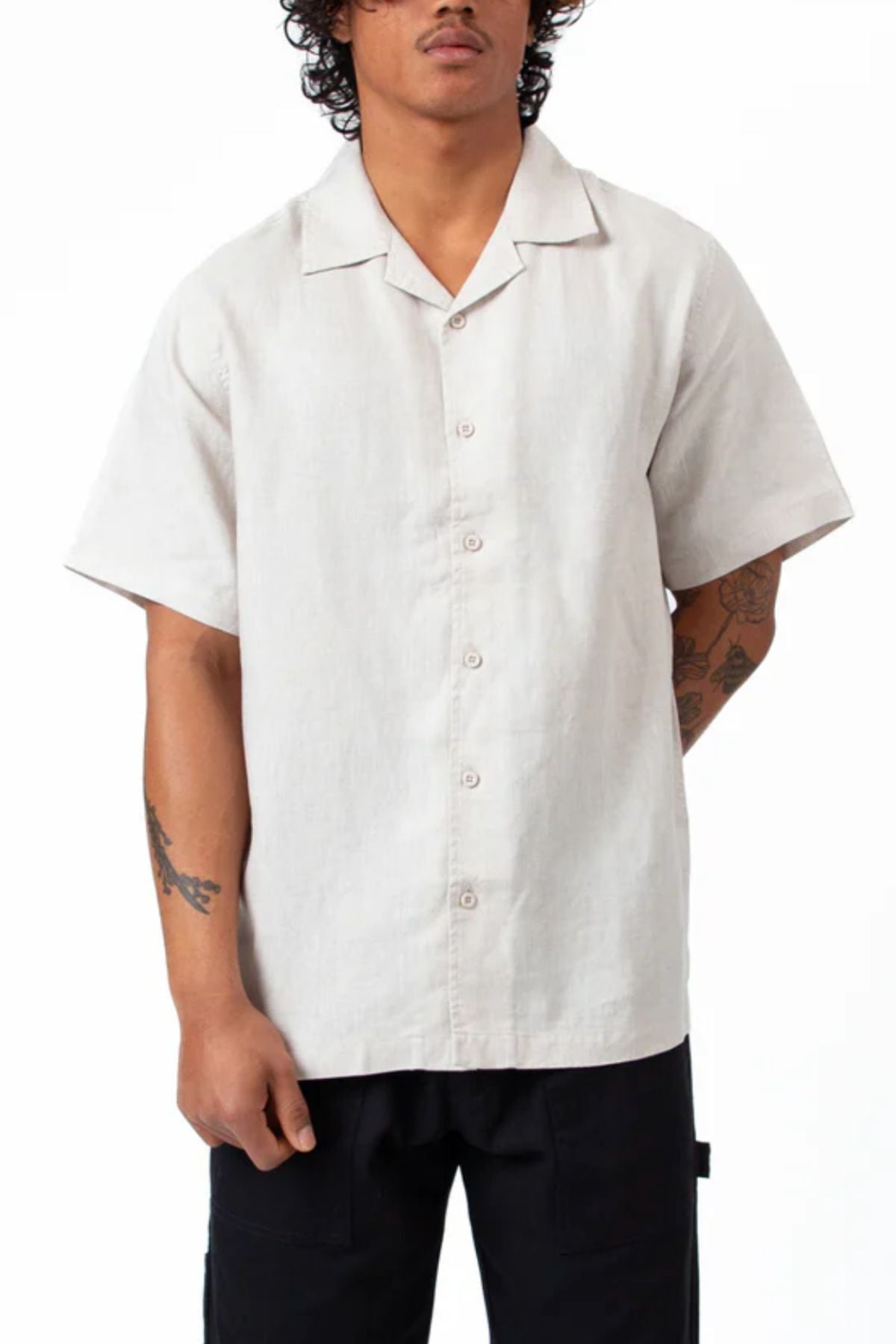 A person with curly hair wears the Mr Simple Cuban Short Sleeve Shirt in light beige and black pants, standing against a white background. Their tattooed arms are relaxed by their sides.