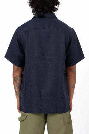 A person with curly hair is seen from behind, wearing the Mr Simple Cuban Short Sleeve Shirt in dark tones and olive green shorts. Arm tattoos are visible, set against a plain white background.