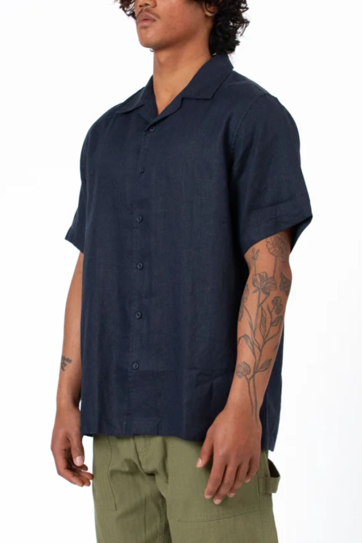 Wearing a relaxed fit, navy blue Mr Simple Cuban Short Sleeve Shirt and olive green pants, a person with floral tattoos on their left forearm stands against a plain white background.