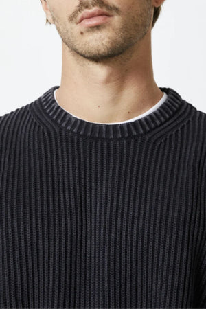 A close-up of a person wearing the Mr Simple Fisher Knit, a black ribbed crew neck jumper layered over a white T-shirt. Only the lower face, neck, and upper chest are shown against a plain light background.