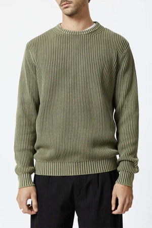 An individual wearing the Mr Simple Fisher Knit sweater in olive green, featuring vertical ribbing, is standing facing forward. The organic cotton sweater boasts a round neckline, long sleeves, and a relaxed fit. The person pairs this with black pants and has a white shirt subtly showing at the neckline.
