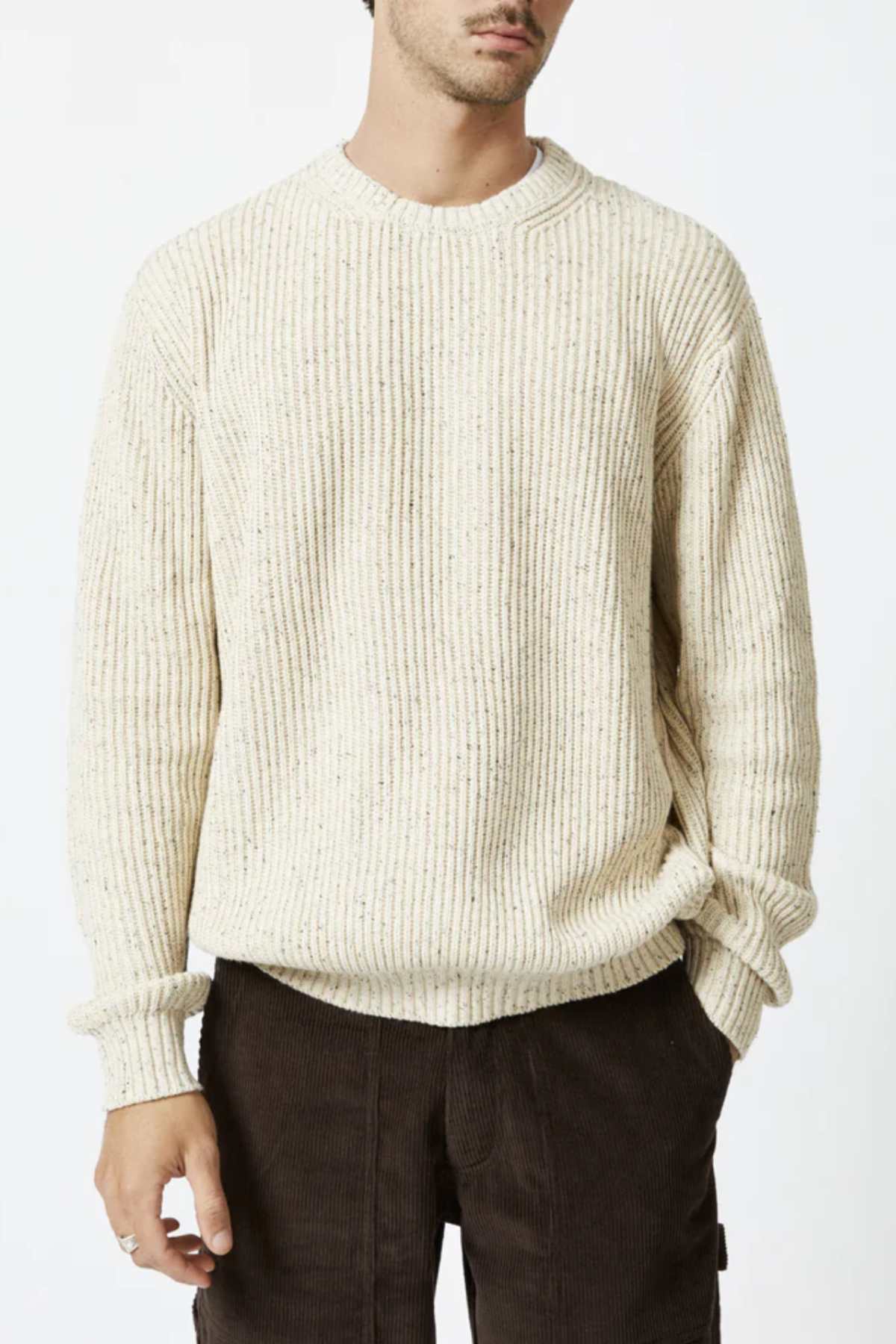 A man stands against a white background, wearing the Mr Simple Fisher Knit—a cream-colored organic cotton sweater with subtle bobble knit and long sleeves—paired with dark brown corduroy pants. His face is partially out of frame.