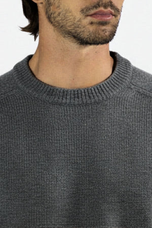 A close-up of a person wearing the Mr Simple Fred Knit—a grey, ribbed, 100% wool jumper with a crew neck. Only the lower face, neck, and upper chest are visible against a plain light background. A timeless addition to your winter wardrobe.