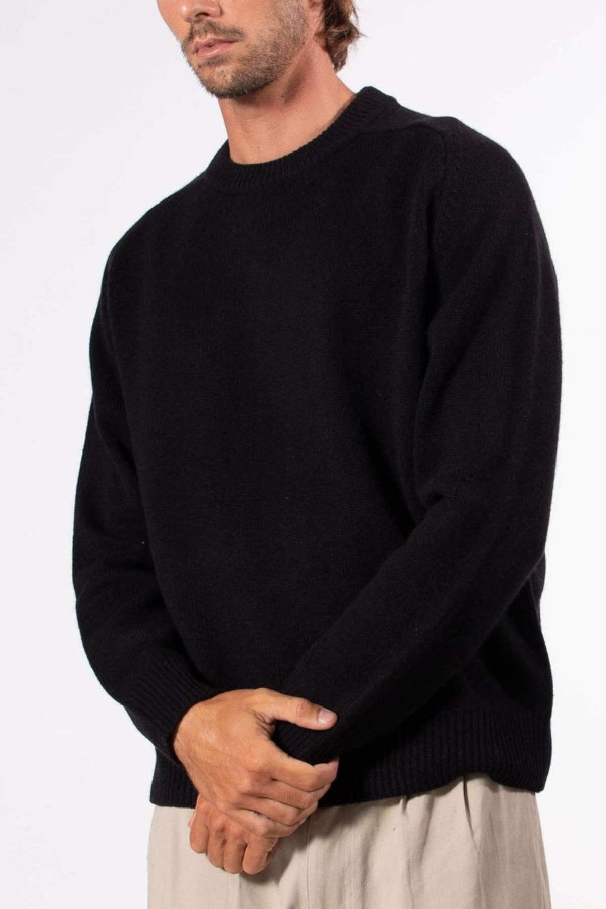 A man wears the Mr Simple Fred Knit, a relaxed fit black wool jumper, paired with light beige pants, standing with hands clasped against a white background—an ideal choice for a stylish winter wardrobe.