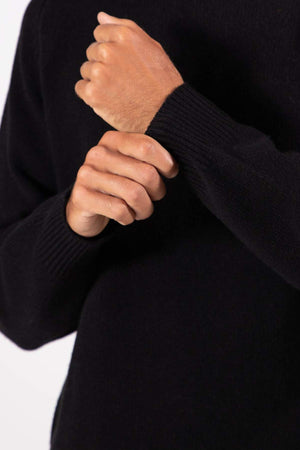 A person wearing the Mr Simple Fred Knit, a black relaxed-fit wool jumper, adjusts the cuff of their left sleeve with their right hand. The image is cropped to display only the upper torso and arms.