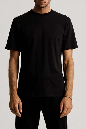 A man stands against a plain background wearing the Mr Simple Heavy Weight Tee, which has a boxy fit and is made from 100% organic cotton, paired with black pants. His arms are relaxed by his sides and his face is partially out of frame.