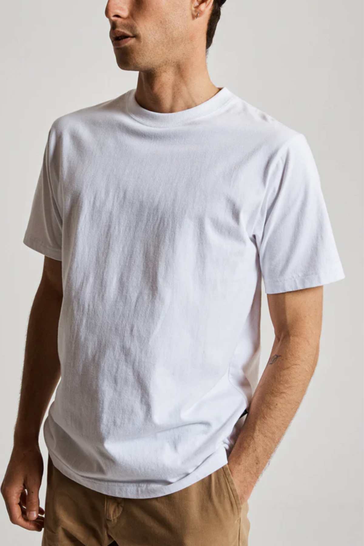 A person in a Mr Simple Heavy Weight Tee (plain white) and light brown pants stands with one hand in their pocket, exuding comfort. Set against a neutral gray background, the focus is on their 100% organic cotton attire, with their face partially obscured.