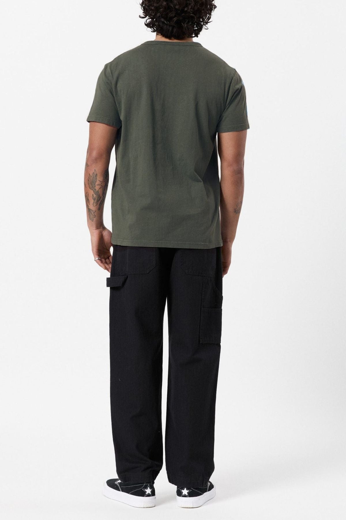 The image features a person standing with their back to the camera, wearing a green Mr Simple Hemp Tee, black cargo pants, and black sneakers with white soles. They have curly hair, a tattoo on their left arm, and are set against a plain white background that highlights sustainability.