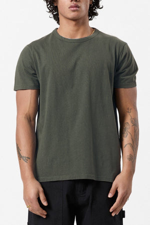 A person in an eco-friendly Mr Simple Hemp Tee in dark green, paired with black pants, stands against a white background. They have curly hair and visible tattoos on both arms, highlighting their commitment to sustainability in fashion.