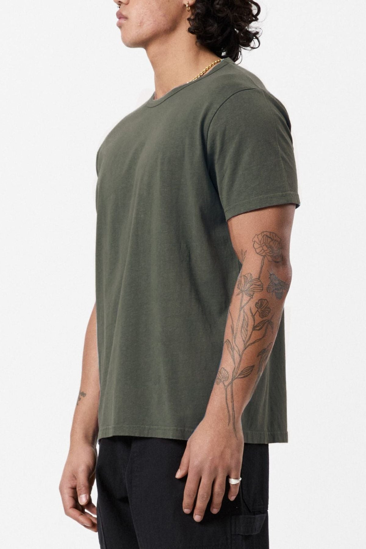 A person with curly hair stands sideways, wearing a Mr Simple Hemp Tee in olive green and black pants. Their arm is adorned with tattoos featuring floral designs, and they are sporting a ring on one finger. The emphasis on sustainability is highlighted against the plain white background.