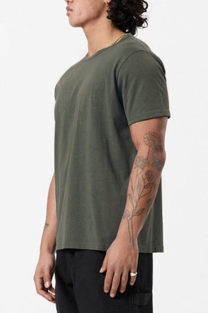A person with curly hair stands sideways, wearing a Mr Simple Hemp Tee in olive green and black pants. Their arm is adorned with tattoos featuring floral designs, and they are sporting a ring on one finger. The emphasis on sustainability is highlighted against the plain white background.