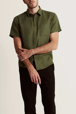 A man wears the Mr Simple Linen Short Sleeve Shirt in olive green, featuring a chest pocket, paired with black pants against a plain light backgroundβan ideal addition to any summer wardrobe. His left hand rests on his right arm.