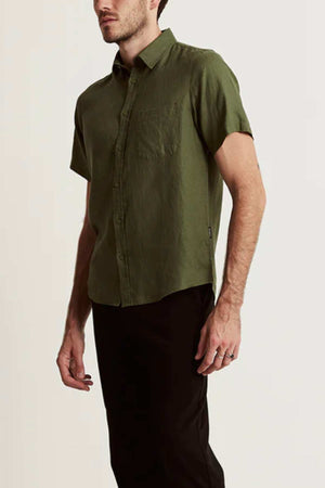 A man wears the Mr Simple Linen Short Sleeve Shirt in olive green with a chest pocket and black pants, standing against a plain light background. The lightweight shirt is ideal for an effortless summer wardrobe.