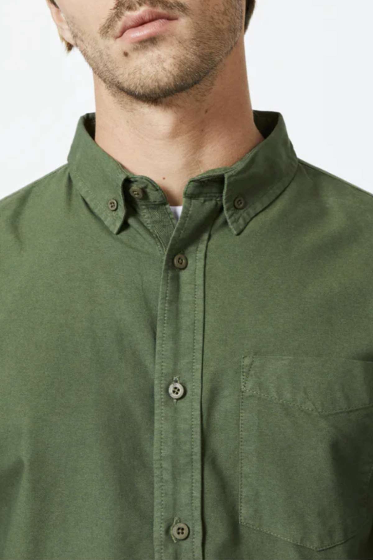 The image features a person in a slim-fit Mr Simple Oxford Shirt, made from organic cotton, showcasing the upper torso. The green button-up includes a left-side pocket, with a hint of facial hair and their face partially visible.