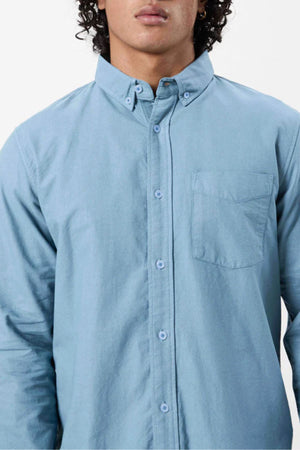 A person wears the Mr Simple Oxford Shirt in light blue, slim-fit with collar and chest pocket, shown from shoulders to waist against a white background. The 100% cotton shirt’s front is partially out of frame.
