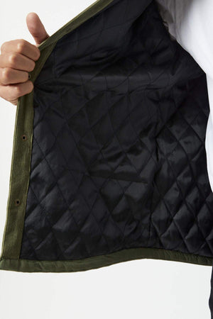 A person holds open the olive green Mr Simple Quilted Cord Jacket, showing its black quilted lining. Part of the jacket is visible as their hand grips the edge near the metal snaps.