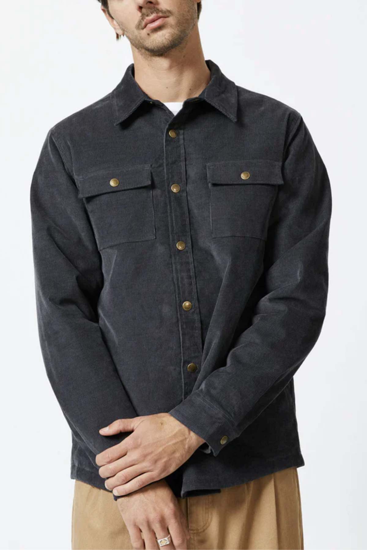 A man wearing the Mr Simple Quilted Cord Jacket, a dark grey button-up jacket with pockets on both sides of the chest. The relaxed fit jacket features brass buttons and is paired with light tan pants. He has one hand touching the opposite wrist and appears to be posing against a plain background.