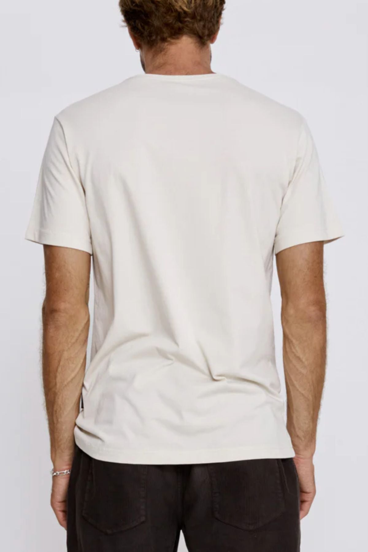 A man with short, light brown hair is seen from behind wearing the Mr Simple Reginald Organic Tee, a plain midweight t-shirt with a comfortable fit, along with dark pants against a neutral background.