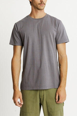 A person in a Mr Simple Reginald Organic Tee paired with olive green pants stands against a white backdrop. The tee, made from organic cotton, offers both comfort and a durable stretchy design. A tattoo adorns their left arm, and their facial expression is neutral.