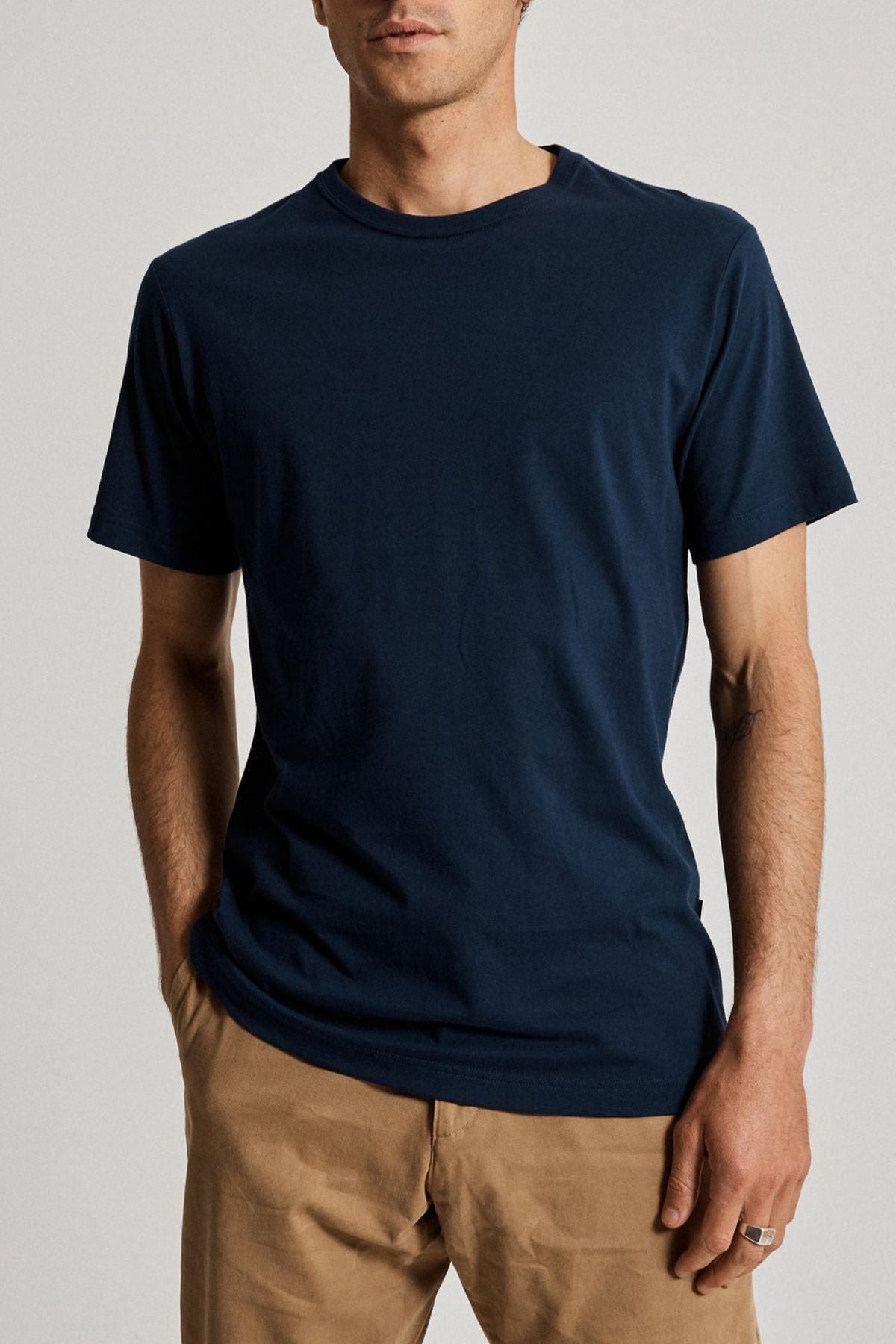 Dressed in a navy Mr Simple Reginald Organic Tee paired with beige pants, an individual stands against a simple background, one hand casually in their pocket. The emphasis is on the relaxed and eco-friendly style of the attire.