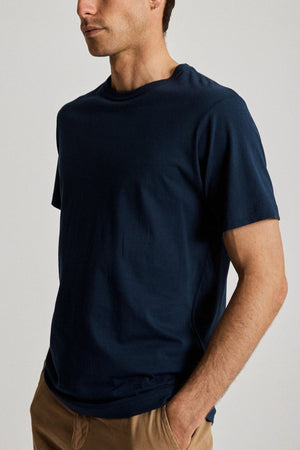 A person wearing the navy blue Mr Simple Reginald Organic Tee, crafted from organic cotton with a sustainable fit and classic design, pairs it with beige pants while standing against a plain, light background. Their arms are relaxed at their sides, with one hand tucked into a pocket.