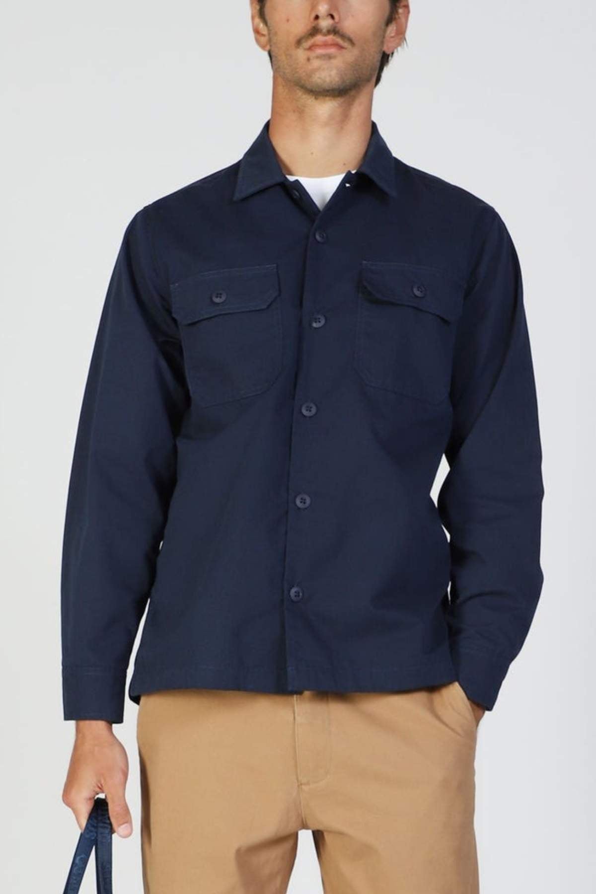A man stands against a plain light background, wearing the Mr Simple Ripstop Canvas Overshirt in navy blue with chest pockets and khaki pants, holding a dark object in his left hand.