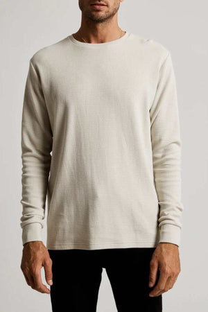 A man wearing a Mr Simple Smith Waffle cream long-sleeved shirt, crafted from organic cotton for a classic fit with a crew neckline, stands against a plain background. He is also in black pants.