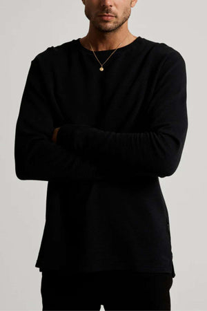 A person in a black Mr Simple Smith Waffle organic cotton long-sleeve shirt and a simple necklace with a small pendant stands against a plain background, arms crossed, with their face not visible.