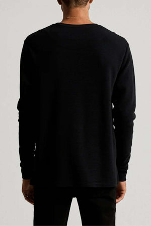 Wearing the Mr Simple Smith Waffle black long-sleeve shirt and matching pants, a person faces away from the camera. The plain, neutral background highlights the textured detail of their organic cotton outfit.
