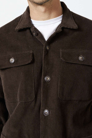 A person is wearing the Mr Simple Stockyard Sherpa Jacket, a dark brown corduroy piece with buttoned pockets over a white shirt, highlighting the texture and full button front design crafted from 100% cotton outer material.