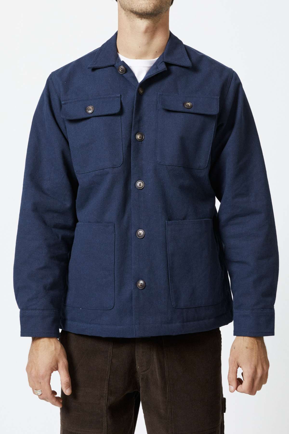An individual is donning a dark blue Mr Simple Stockyard Sherpa Jacket, featuring buttoned chest pockets and hand pockets at the waist. The sherpa lining provides added warmth, while the 100% cotton fabric offers comfort. With their hands resting by their sides, they stand against a white background and pair the jacket with brown pants. Their head is not visible in the frame.