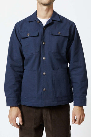 An individual is donning a dark blue Mr Simple Stockyard Sherpa Jacket, featuring buttoned chest pockets and hand pockets at the waist. The sherpa lining provides added warmth, while the 100% cotton fabric offers comfort. With their hands resting by their sides, they stand against a white background and pair the jacket with brown pants. Their head is not visible in the frame.