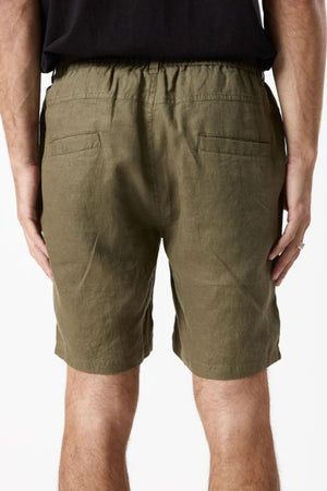 A person wearing Mr Simple Tanner 3.0 Linen Short in breathable olive green with an elastic waistband and two back pockets, paired with a black shirt, shown from the back.