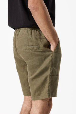 A person in olive green Mr Simple Tanner 3.0 Linen Shorts featuring an elastic waistband and side pockets, seen sideways with one hand in a pocket, paired with a black shirt.