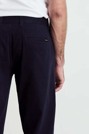 A person in Mr Simple Wilson Standard Chino pants and a relaxed-fit shirt made from organic cotton is shown from behind. Their left arm reveals a small tattoo near the wrist against the plain white background.