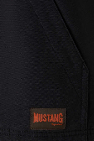 Close-up of a Mustang Signature Outback Canvas Vest made from durable canvas material, featuring a dark fabric with a brown patch label at the bottom. The label reads "MUSTANG Signature" in bold orange and cursive red lettering. The fabric has a seam running diagonally across the image, highlighting its functional features.