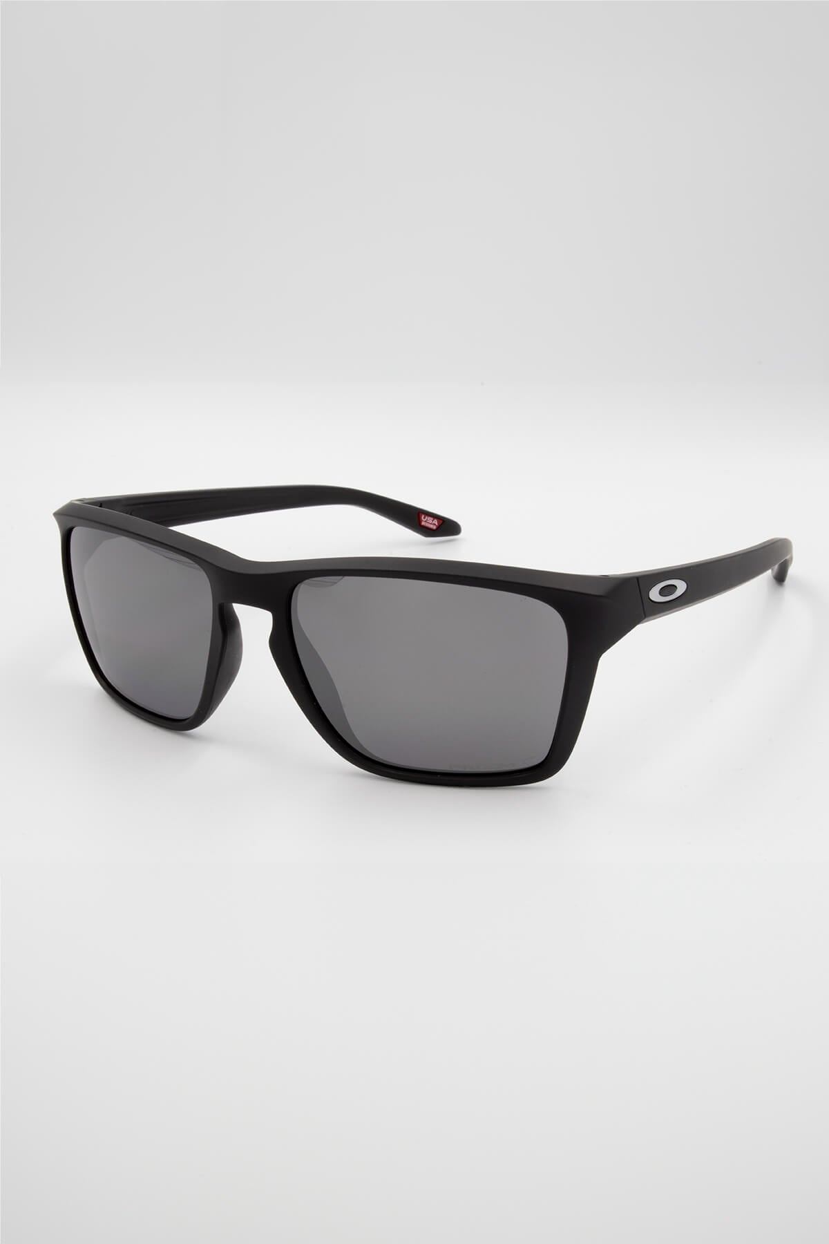 A pair of sleek, black Oakley Sylas Polarized sunglasses featuring a rectangular frame and dark grey lenses. The right temple showcases a small white "O" logo. Enhanced with Prizm lens technology, these sunglasses are positioned at an angle on a plain white background.